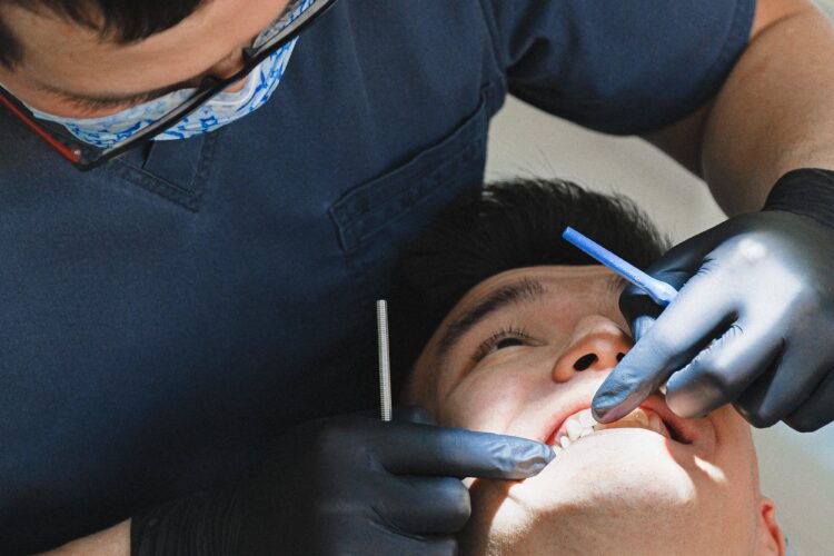 Dentist in a navy blue uniform and black gloves performing a dental procedure on a reclined patient with their eyes closed. The dentist is using an instrument and a suction tool in the patient's open mouth.