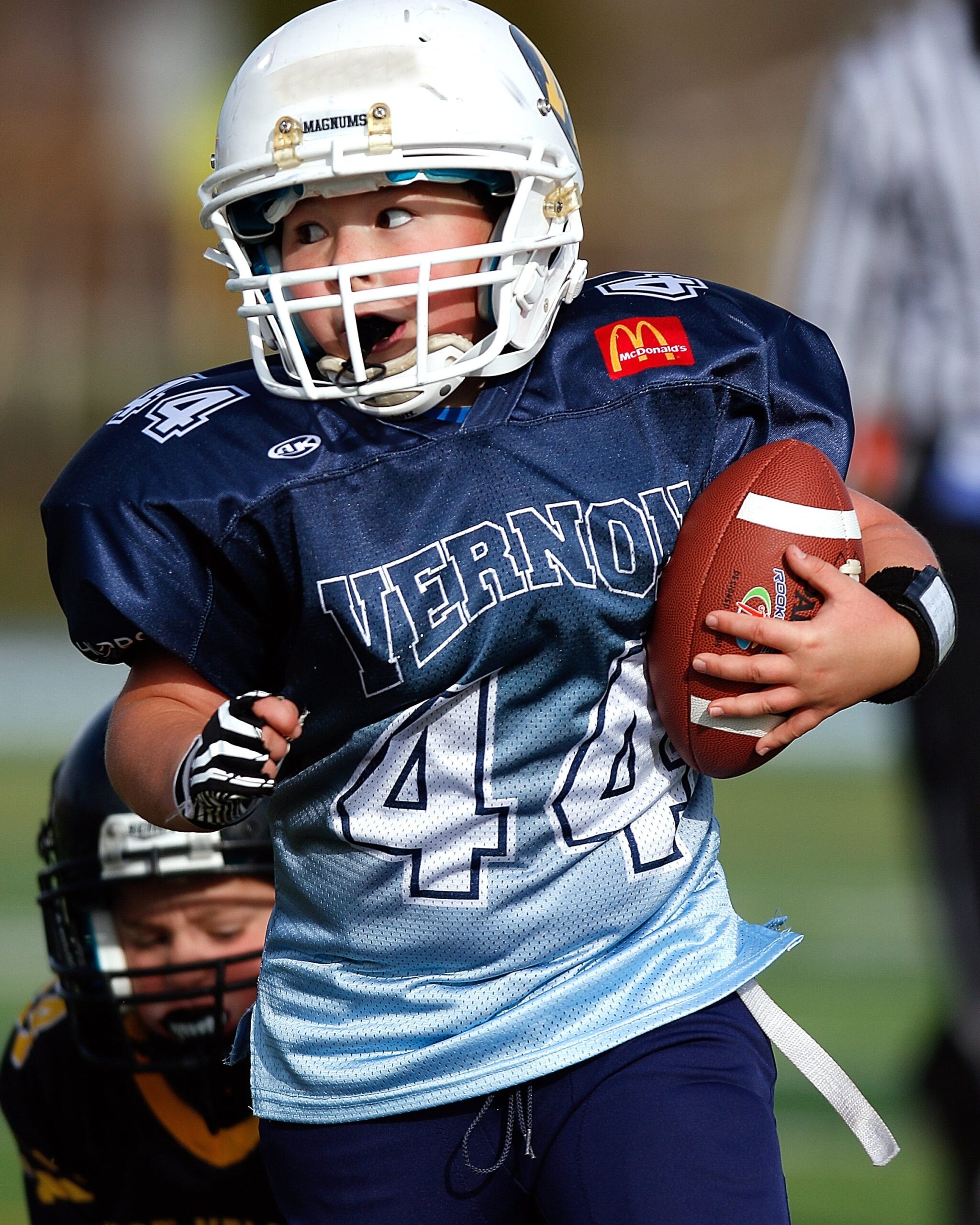 A child in a navy blue football jersey and a white helmet running with a football, looking ahead attentively during a game