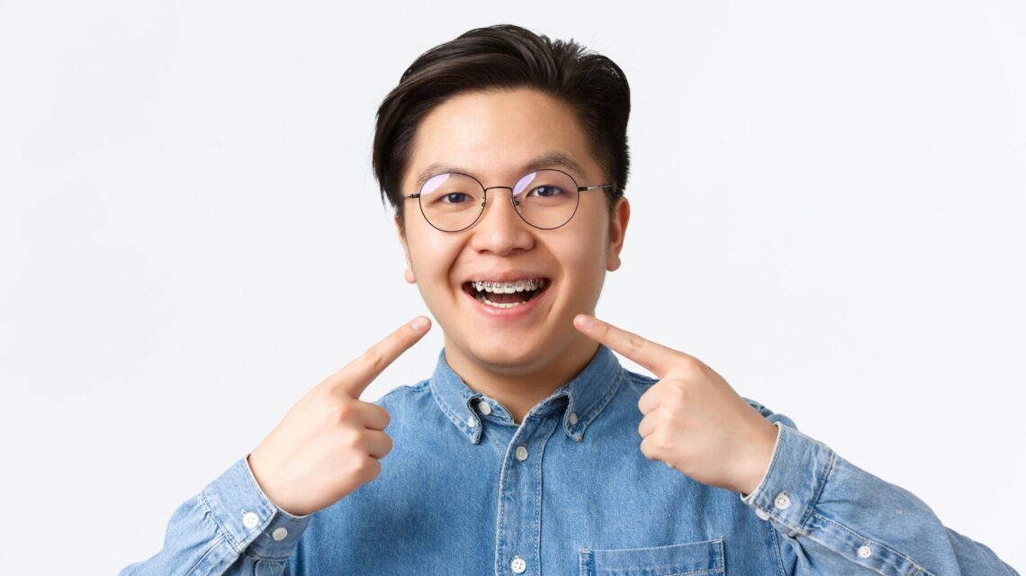 Close-up of satisfied asian guy, dental clinic client smiling happy and pointing at his dental braces, standing white background, recommend quality