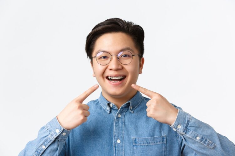 Close-up of satisfied asian guy, dental clinic client smiling happy and pointing at his dental braces, standing white background, recommend quality