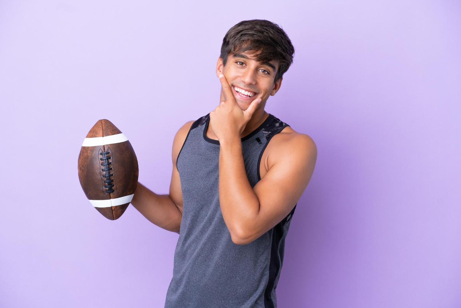 Smiling young man holding a football and touching his chin against a purple background.
