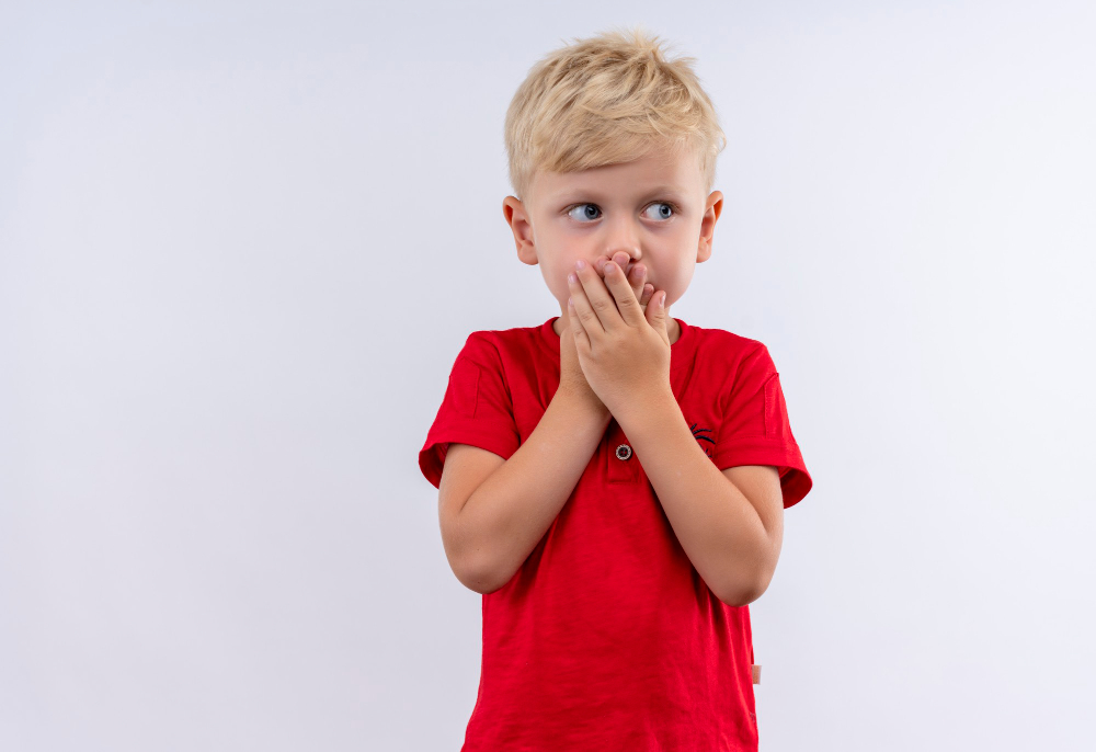 A surprised little cute blonde boy in red t-shirt keeping hands on his mouth while looking side on a white wall